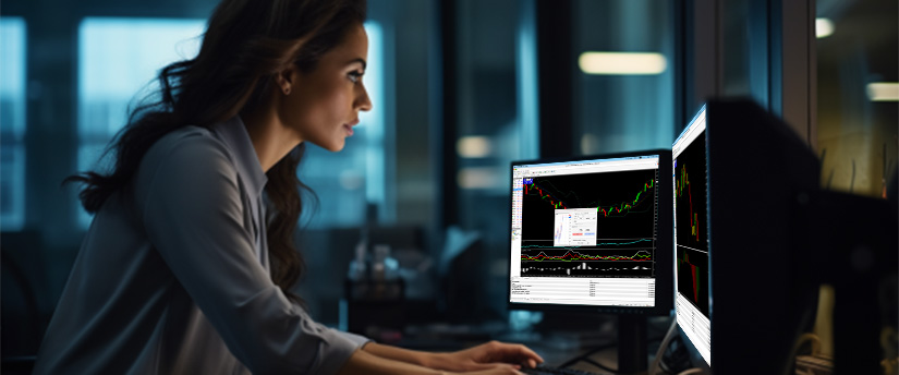 Uma mulher trabalhando em uma mesa com dois monitores, cada um exibindo diferentes gráficos de tendências do mercado de ações.