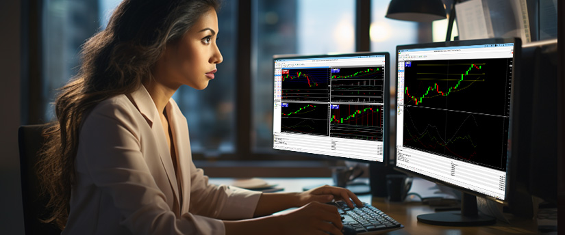A woman at a desk with two monitors displaying different forex stock market graphs.